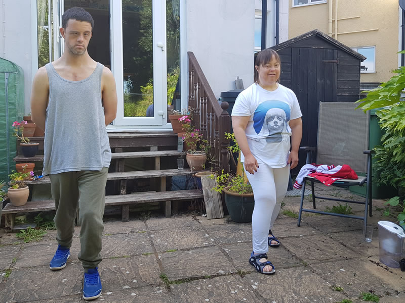 Two adults walking across a paved back garden on a summer day, past potted plants, wooden steps and a folding chair